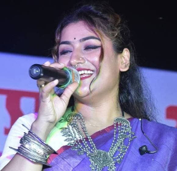Arpita Chakraborty singing into a handheld microphone, laughing mid-phrase, in a purple sari and heavy silver jewellery