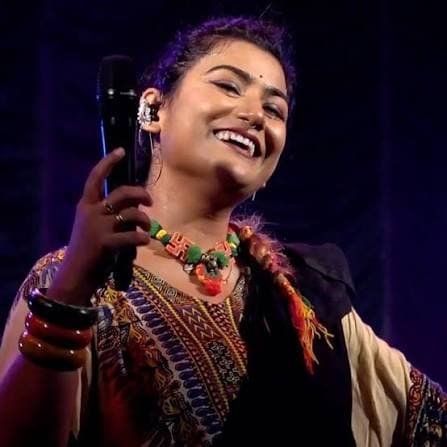 Arpita singing on stage, microphone in hand, wearing a beaded folk necklace over an embroidered kurta