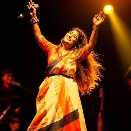 Arpita on stage with both arms flung up, hair loose, lit by a single warm spotlight