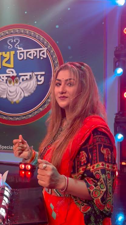 Arpita on a television set in a red sari, in front of the show’s illuminated Bengali signage